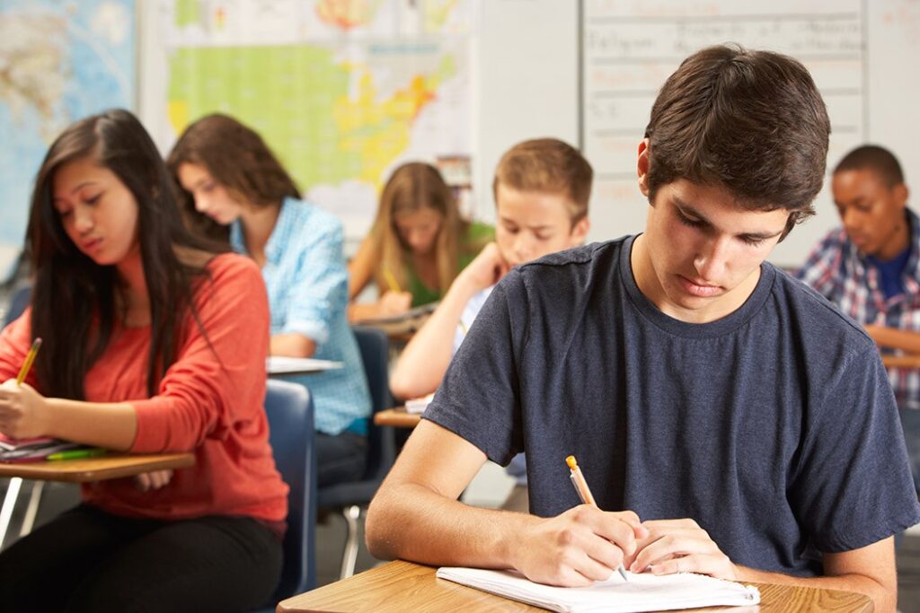 A classroom full of studious high school students with a young while male student writing in the foreground